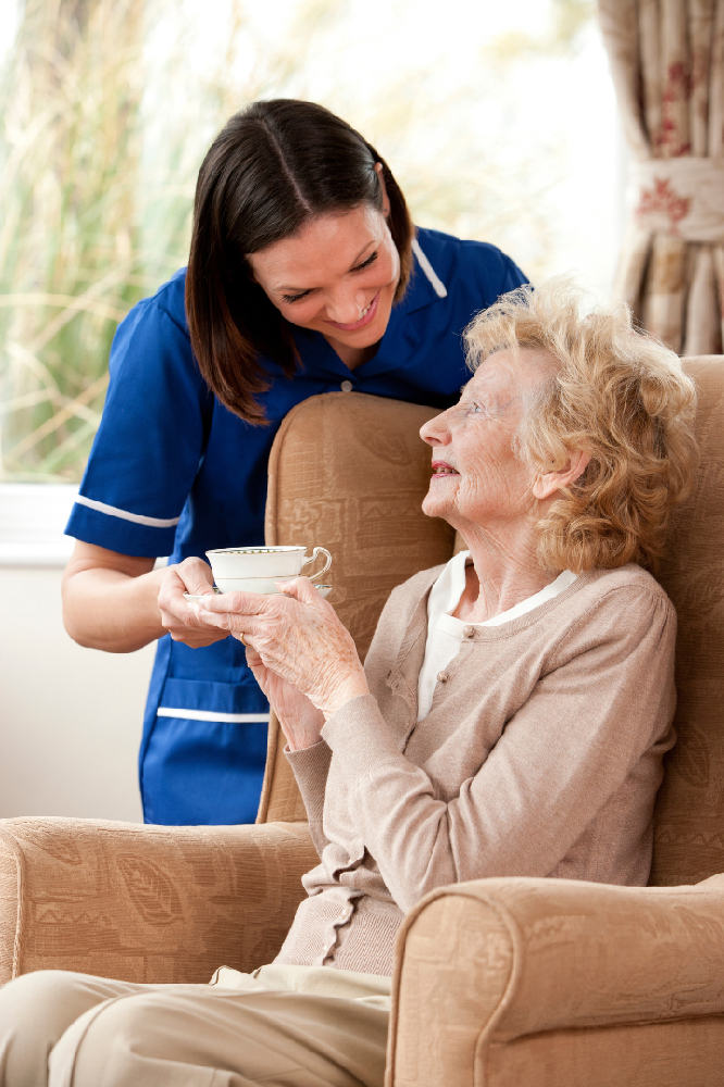female care assistant handing an elderly lady a cup of tea whilst she sits in an armchair